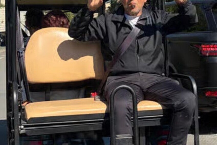 A person sitting on a tan bench with a black jacket, a black car in the background, and a red object on the bench. 5