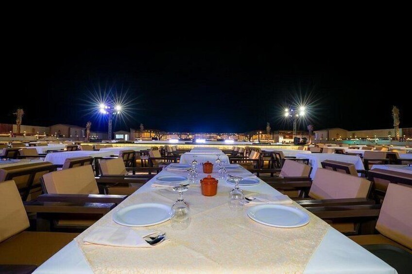 An outdoor dining area with tables and chairs, a long table with plates and glasses, and a view of the sea in the background. 2