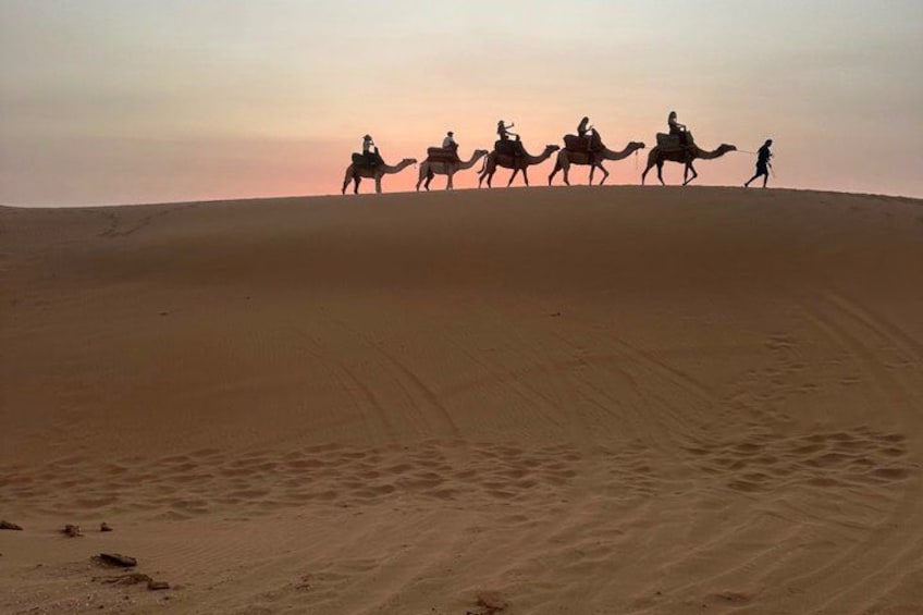 Un groupe de personnes chevauche des chameaux à travers un paysage de dunes de sable, au coucher du soleil. 2