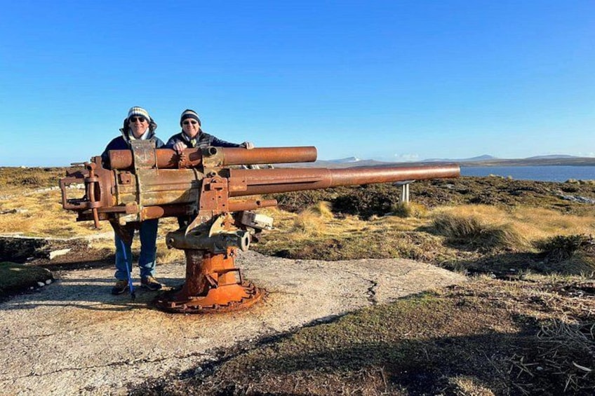 Two people standing next to a large, rusted cannon on a rocky terrain with a clear sky and distant water body. 5