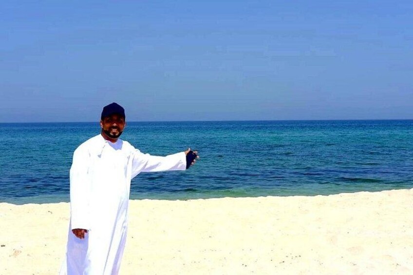 A person in traditional attire standing on a sandy beach with a clear blue sea in the background. 4