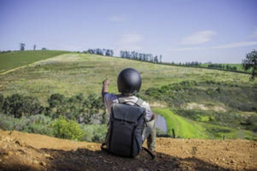 A person sitting on a dirt path, looking at a green field with a hill in the distance. 1