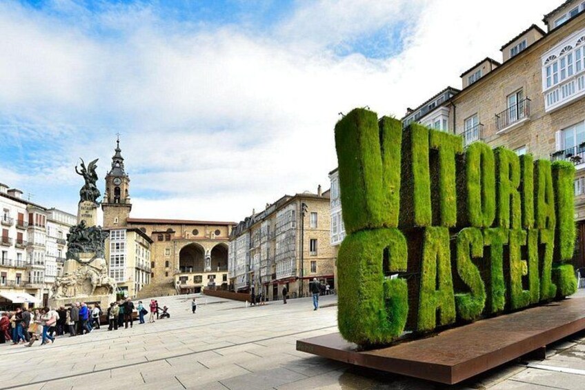 En offentlig plads med en stor, grøn skulptur, der staver 'VITORIA CASTELLO', og en historisk bygning med en statue i baggrunden. 4