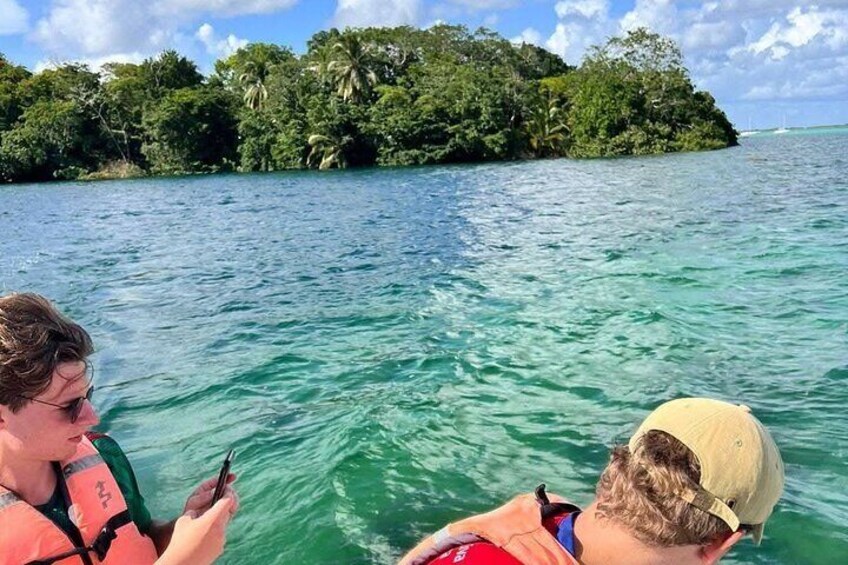 Un bote con dos personas usando chalecos salvavidas, una sosteniendo un teléfono, y una isla verde con vegetación densa al fondo. 4