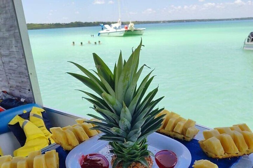 Una piña en un plato junto a fruta cortada y una bebida, vista desde un bote con vista al océano y otro bote a lo lejos. 2