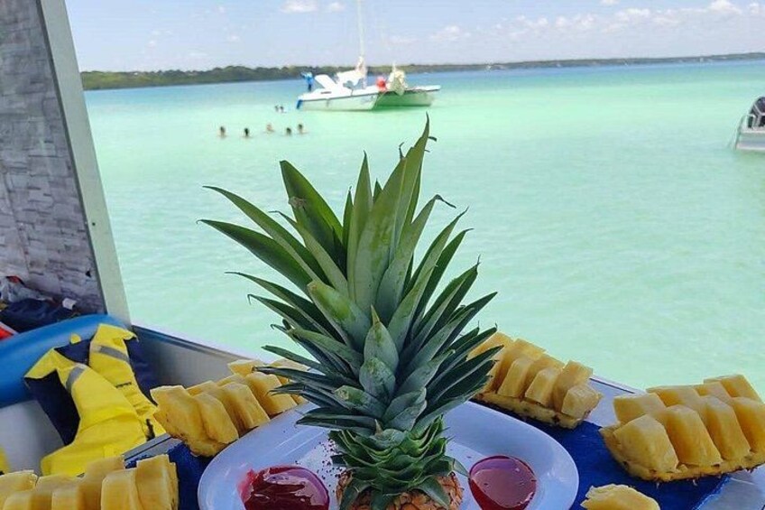 Un centro de mesa con piña, fruta cortada y una bebida sobre una mesa, con vista al agua turquesa y barcos. 2