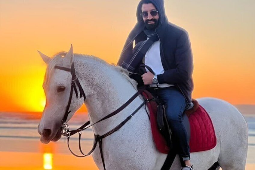 Eine Person reitet auf einem weißen Pferd mit Sattel in Rot am Strand bei Sonnenuntergang. 4