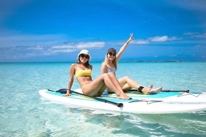 Two people sitting on a surfboard in clear turquoise water with a blue sky. 1