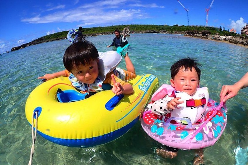 Two children on inflatable rings in clear water with a distant shoreline and wind turbines. 2