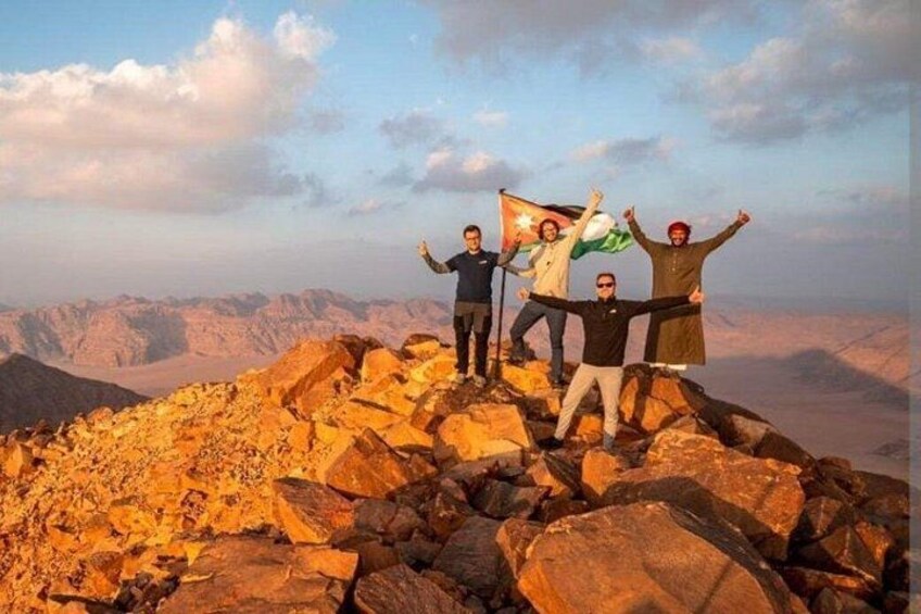 Un groupe de personnes se tient sur un sommet rocheux, un drapeau flotte, et des montagnes se dressent à l'arrière-plan. 1