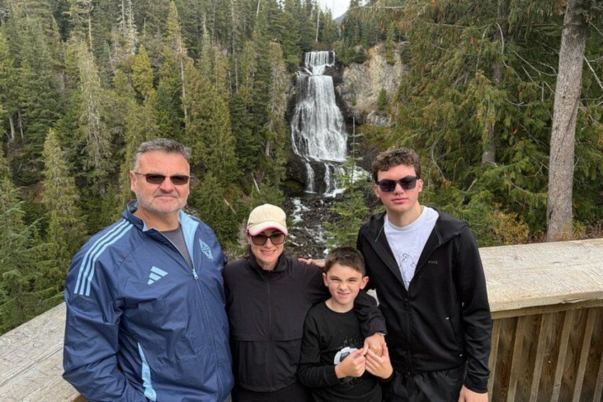 A group of people standing on a wooden deck with a waterfall and forest in the background. 2