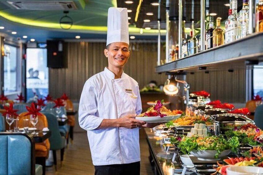 Uno chef con divisa bianca e cappello da cuoco, tiene in mano un piatto, in piedi in un ristorante con un tavolo per buffet ricco di diverse portate e un'area bar con bottiglie. 2