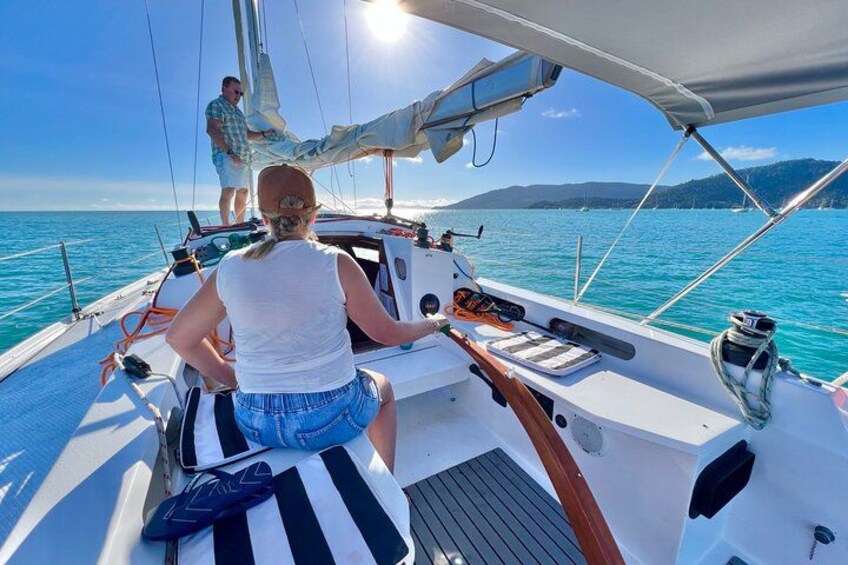 A sailboat on a calm sea with a person steering and another standing at the stern. 2