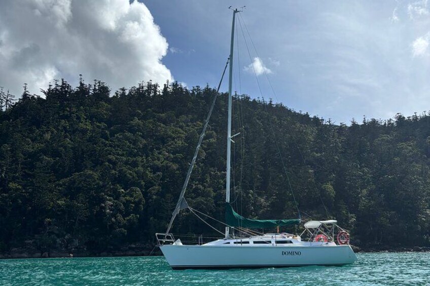 A sailboat named 'DOMINO' on calm turquoise waters with a forested island in the background. 3