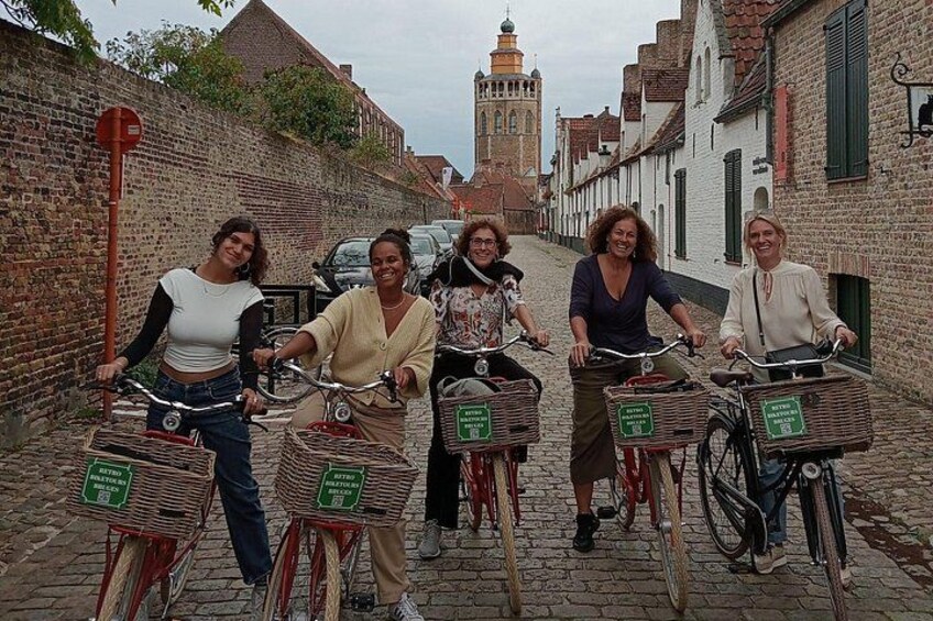 En grupp människor cyklar på en kullerstensgata. De har korgar monterade frampå cyklarna. På ena sidan av gatan finns en tegelvägg, och i bakgrunden skymtar ett torn. 2