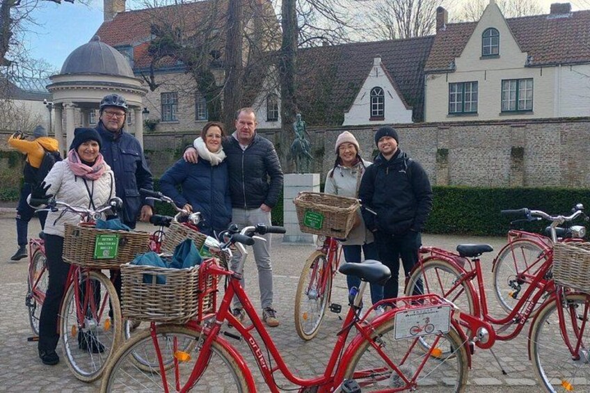 En grupp människor som står framför röda cyklar med korgar, på en kullerstensbelagd plats med historiska byggnader i bakgrunden. 4