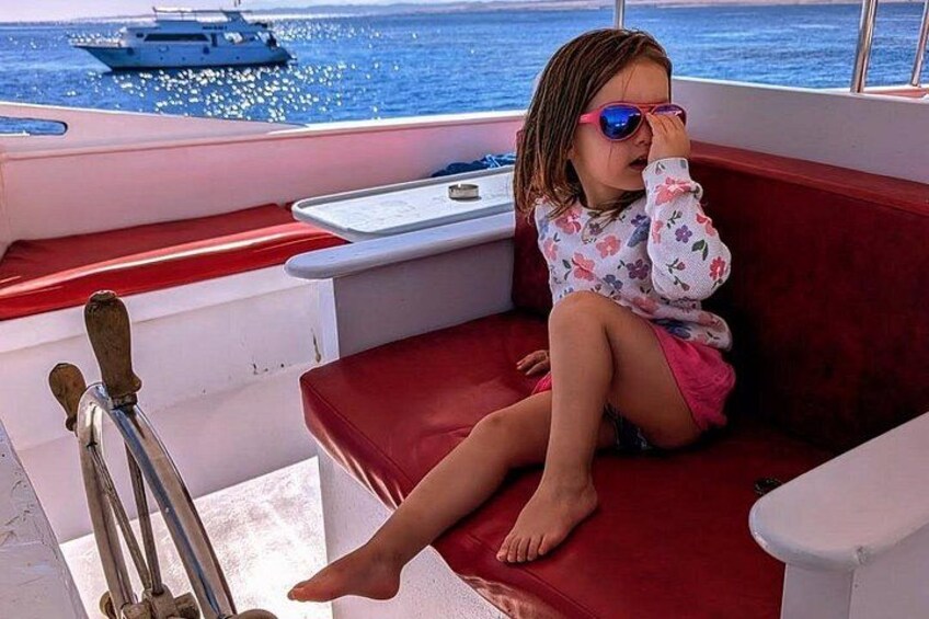 A child sitting on a red bench on a boat, with water and a distant ship in the background. 5