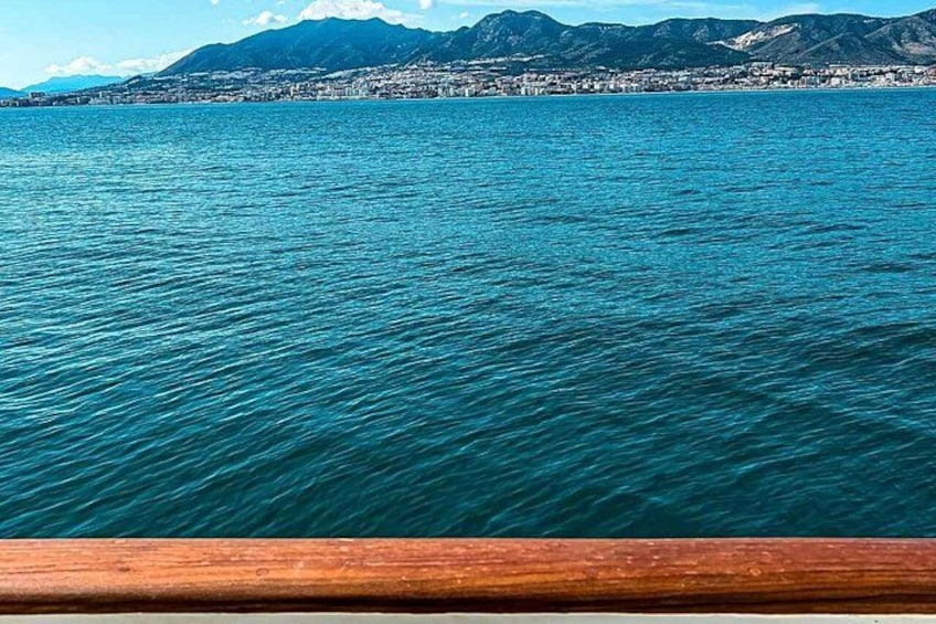 A body of water with a wooden railing in the foreground, a distant shoreline with buildings, and mountains in the background. 2