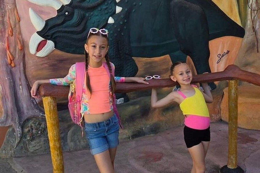 Two children standing on a wooden beam in front of a dinosaur mural. 2