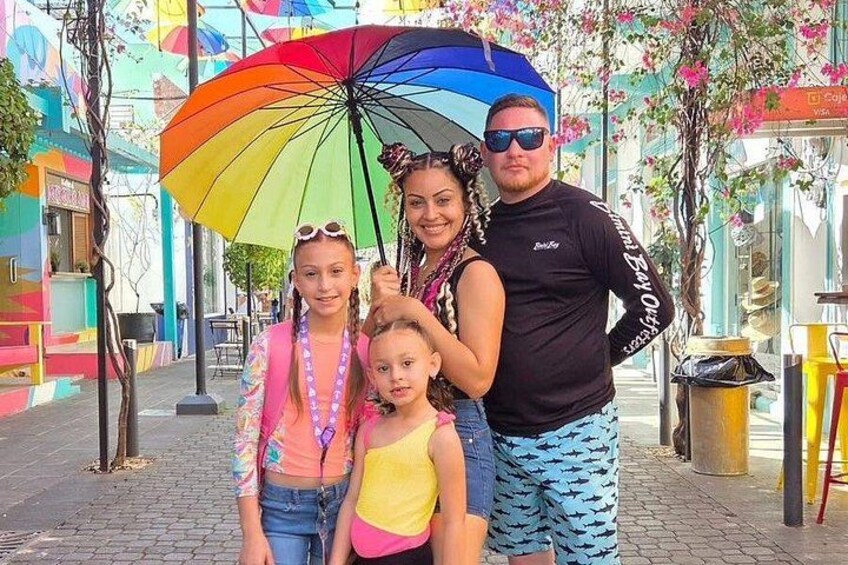 A family with a colorful umbrella, a street with colorful buildings, and a trash can. 5