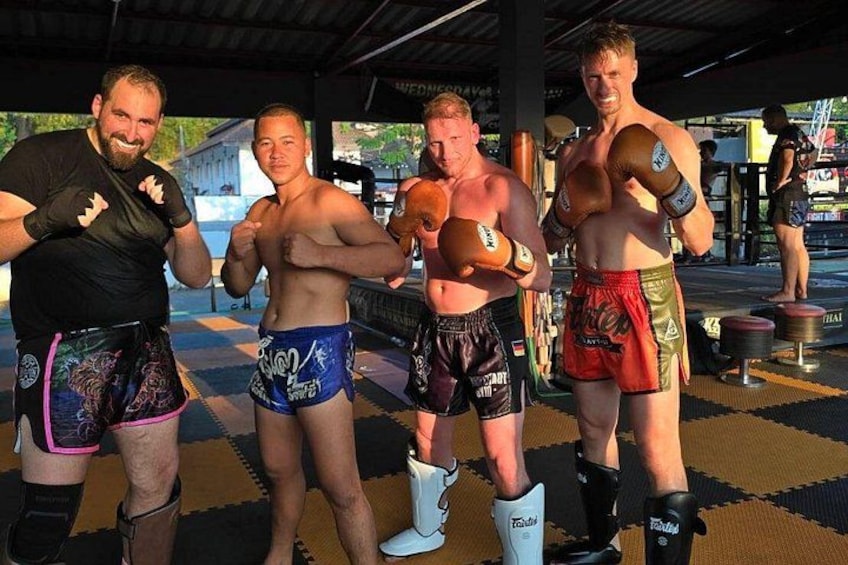 Four individuals in a gym setting, wearing boxing gloves and shorts, with a checkered floor and equipment in the background. 5