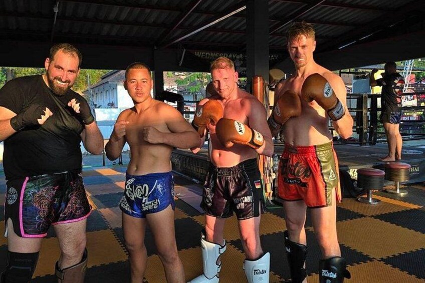 Four men in athletic wear, standing on a checkered mat, with a covered structure and a person in the background. 2