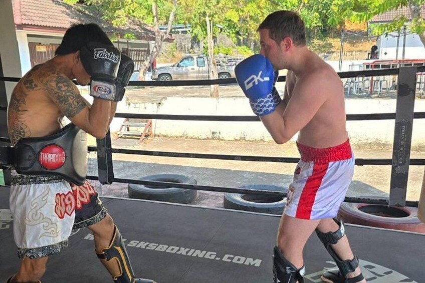 Two people in a boxing ring, one wearing red and white shorts and the other in black shorts, with a tire and a building in the background. 4