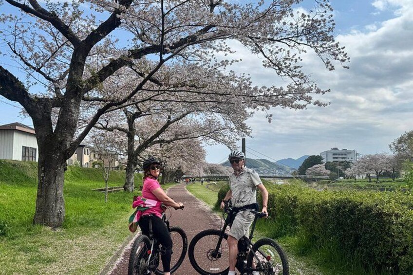 Two cyclists on a path with cherry blossoms, a building, and a hilly landscape in the background. 4