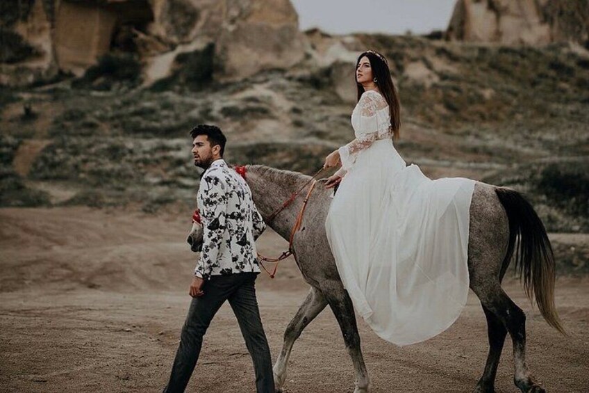 A person in a white dress riding a horse, another person walking beside the horse, and a rocky desert landscape in the background. 4