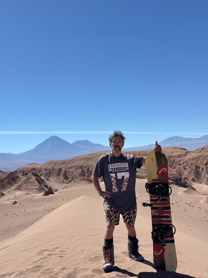 A person standing on a sandy beach with a snowboard leaning against them, mountains in the background. 4