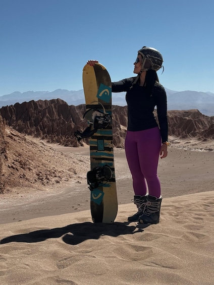 A person holding a snowboard with a camera attached, standing on a sandy terrain with mountains in the background. 1