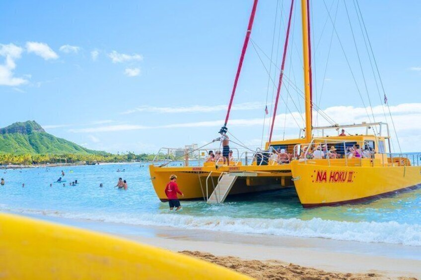 Un bote amarillo llamado 'NA HOKU III' en una playa con gente nadando y una montaña al fondo. 4