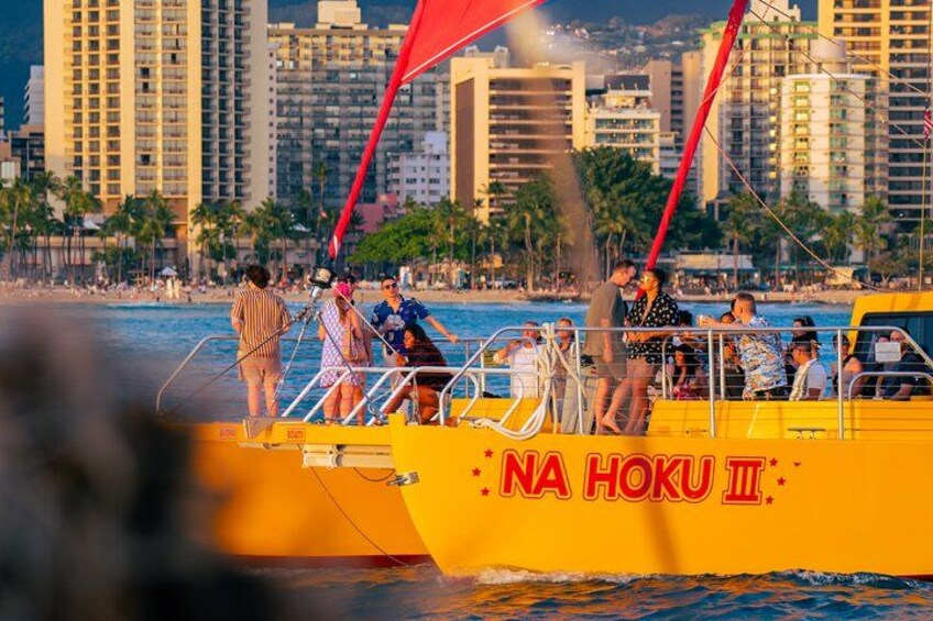 Un bote amarillo con la inscripción 'NA HOKU III', amarrado en una playa con palmeras y edificios al fondo. 3