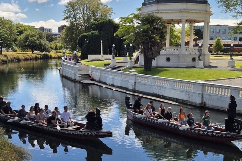 A group of people in a rowboat on a calm river, with a gazebo and trees in the background. 3