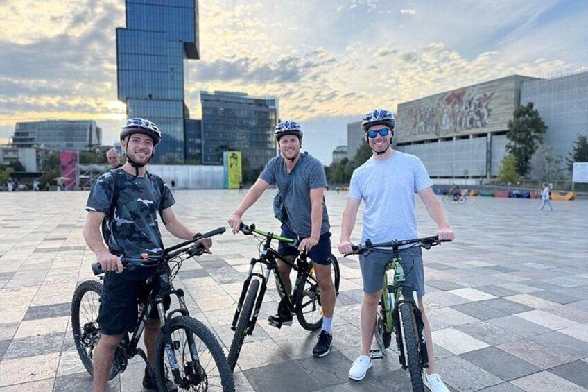 Tres personas en bicicleta en una plaza urbana con edificios modernos al fondo. 4