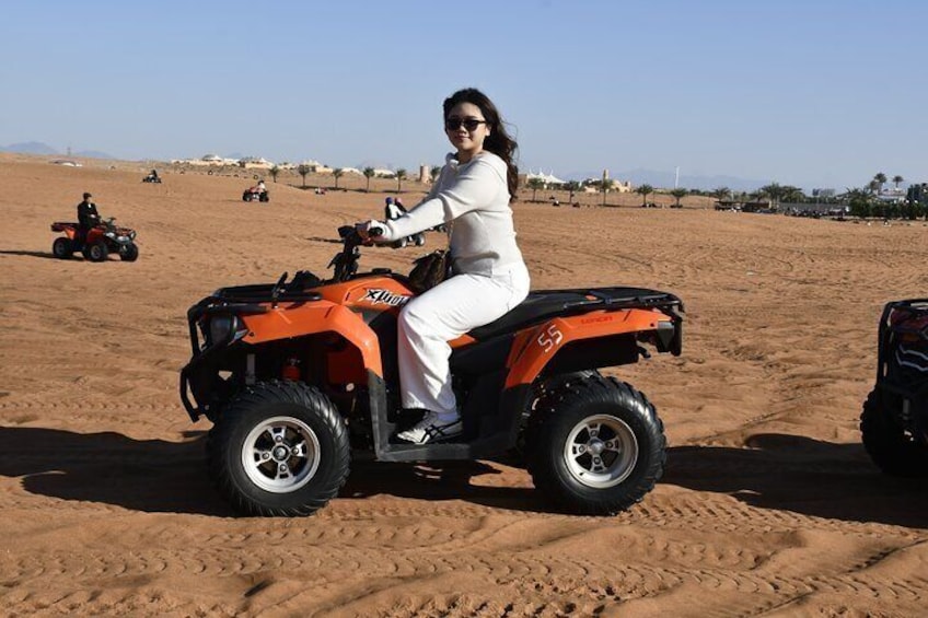 A person riding an orange ATV in a desert with sand dunes and other ATVs in the background. 2