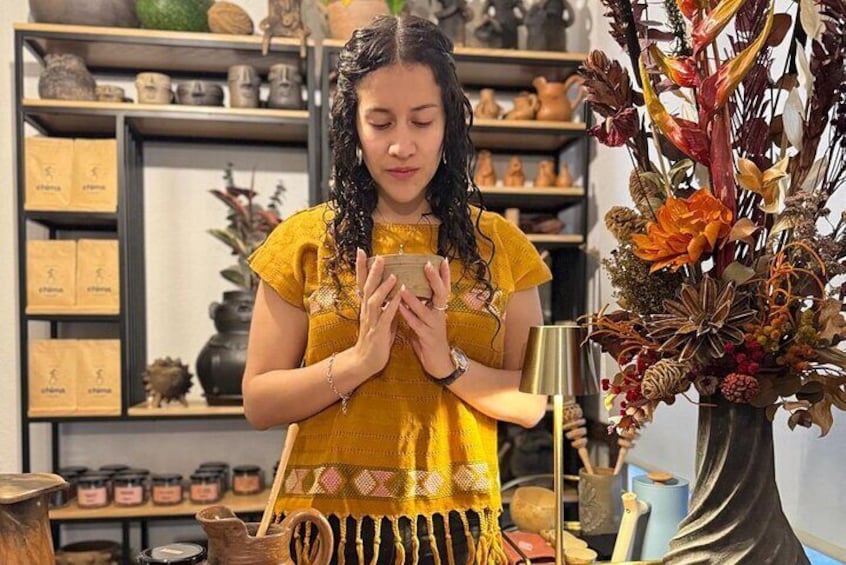Une personne vêtue d’une robe jaune tient un petit objet dans ses mains, et elle est entourée d’étagères remplies de poteries et d’un vase décoratif contenant des plantes séchées. 1