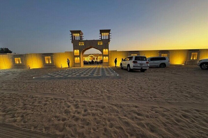 A sandy area with a large building and two towers, surrounded by parked cars. 2