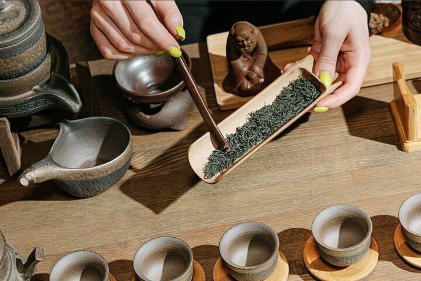 A person holding a wooden tray with tea leaves, a teapot, and several cups on a wooden table. 4