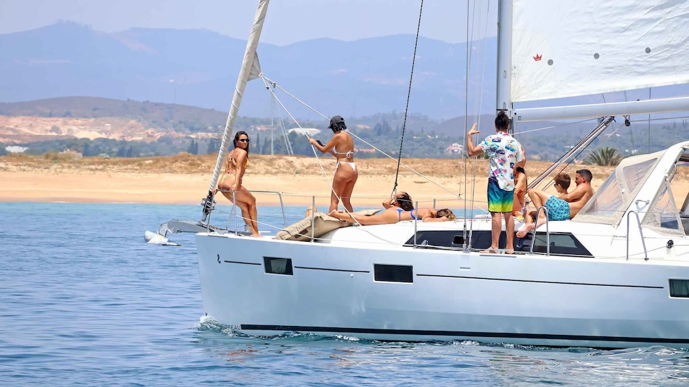 A sailboat on calm waters with people on deck, a sandy beach, and mountains in the background. 5