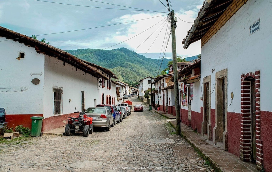 A cobblestone street lined with white and red buildings, parked cars, and a mountainous backdrop. 4