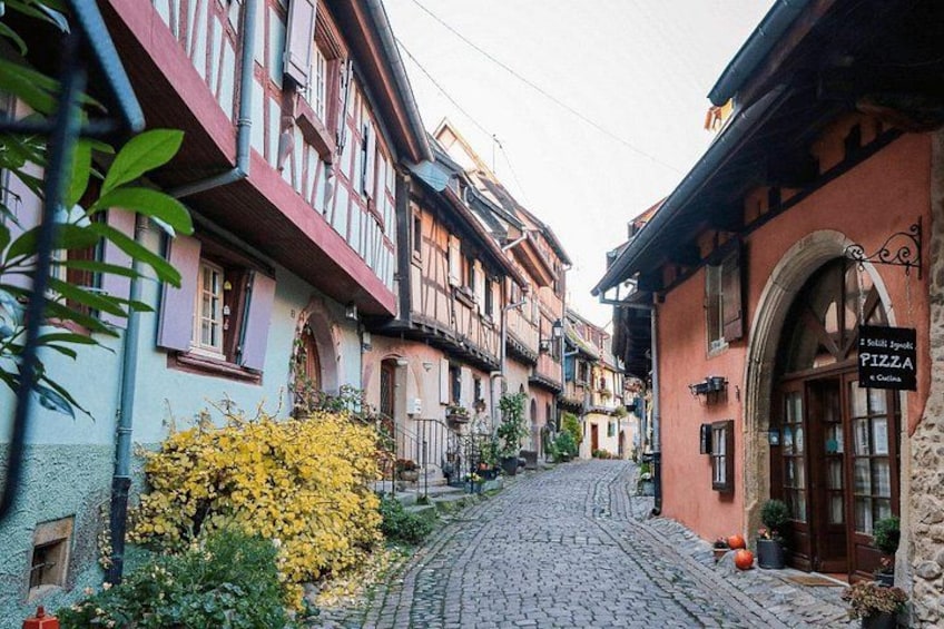 Una calle adoquinada flanqueada por edificios tradicionales con entramado de madera, un letrero de restaurante de pizza y un arbusto amarillo con flores. 1