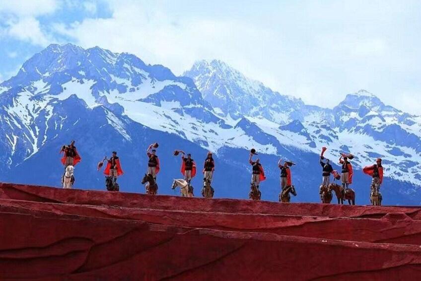 A group of people performing a traditional dance on a red platform with snow-capped mountains in the background. 2