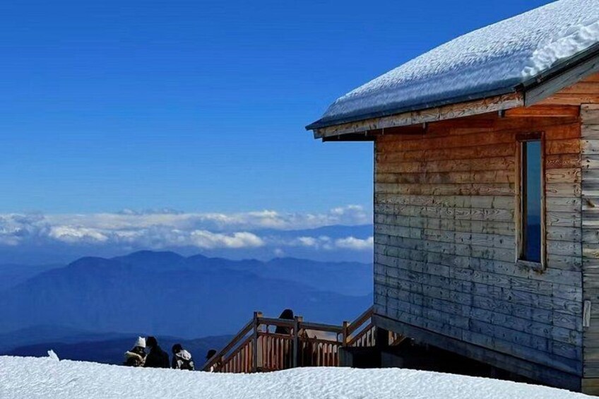 A wooden cabin with a snow-covered roof, a wooden staircase, and a view of distant mountains under a clear blue sky. 3