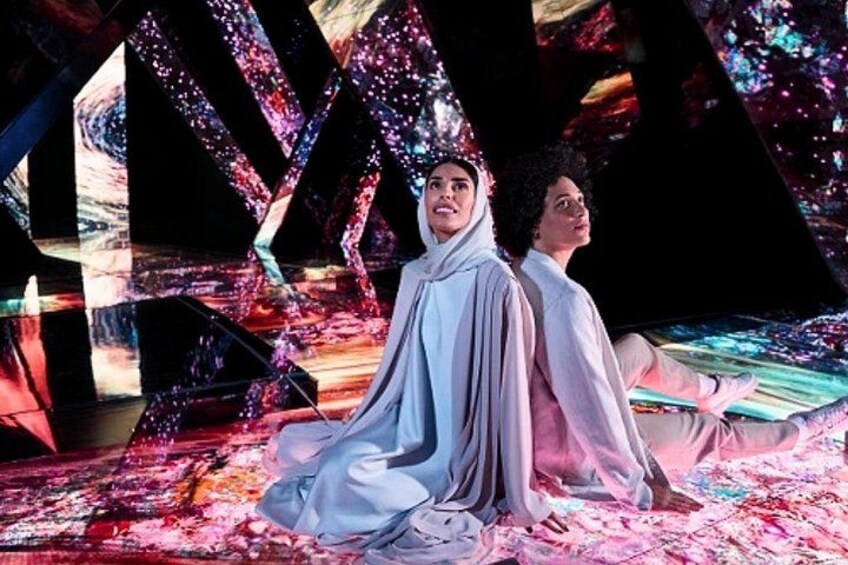 Two people in flowing white garments seated on a colorful, abstract patterned floor. 4