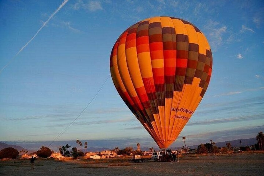 Een luchtballon met een geruit patroon, voorzien van de tekst 'BALLOONBEE BUCKETS', boven een landschap met bomen en een heldere lucht. 1