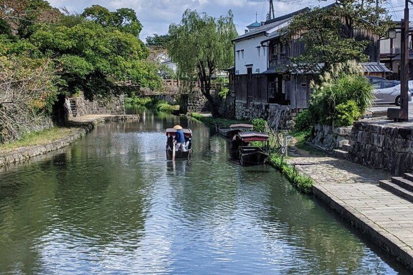 Un étroit canal où navigue un bateau transportant une personne, le tout étant bordé de bâtiments traditionnels et de verdure. 1