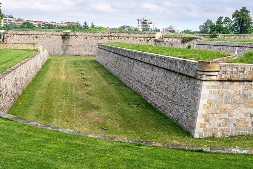 A grassy area enclosed by stone walls with a cityscape in the background. 2