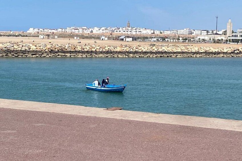A calm body of water with a small blue boat carrying two people, a rocky shoreline, and a distant cityscape. 3