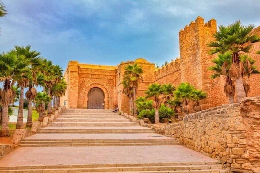 A stone staircase leading to a large arched doorway, flanked by palm trees and a stone wall. 1